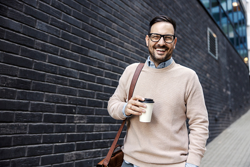 An executive in smart casual is holding takeaway coffee and smiling at the camera.