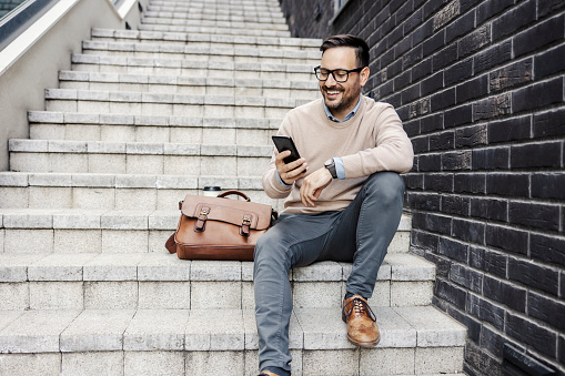 Smart casual businessman is sitting on the stairs outside, holding phone and smiling at it.