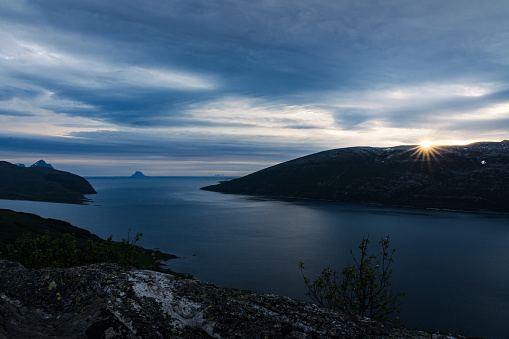 View towards the ocean from a view point high above a fjord in Norway with sun and sun rays above the mountain ridge