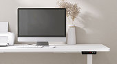 Modern design white height adjustable standing desk with digital controller, computer desktop, printer, flower vase in sunlight on cream wallpaper wall