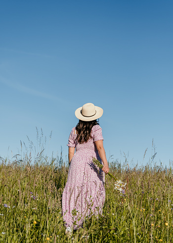 Lifestyle image of beautiful woman with long hair, walking in field or meadow, holding wildflowers.