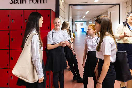 A three-quarter length shot of a group of teenage students and their teacher standing in a hallway in school smiling and talking. They are waiting to go into a class in a school based in the Northeast of England.