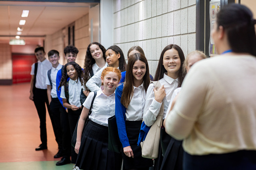 Over-the-shoulder three-quarter-length shot of a group of students lining up for a class at school. They are smiling at an unrecognisable teacher. The school is based in the North East of England.