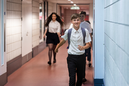 A three-quarter length shot of a male student running through a corridor in school to get to class on time. He is smiling wearing a backpack on his back. The school is based in the North East of England