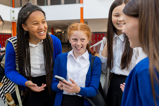 A waist-up shot of a group of female teenage students in their school. They are smiling and talking to each other about something on a mobile phone. This school is based in the North East of England.
