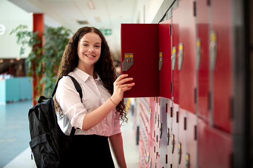 A three-quarter length shot of a teenager opening her locker at school. She is smiling into the camera wearing a full school uniform.