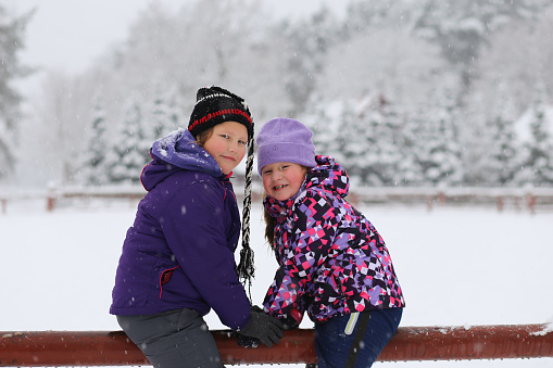 Two smiling laughing girls sisters siblings sitting on farm fence under snowfall. Farm in the pine fir tree forest/ Witer activities sports and leisure