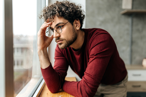 Portrait of depressed man contemplating his thoughts while standing next to the window in the apartment