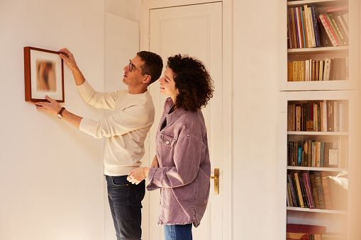 A young couple standing and holding a painting in front of a wall.