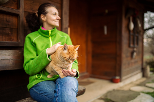 Mature woman relaxing in front of cottage at countryside, sitting cozy on bench while her cat sitting in her lap.