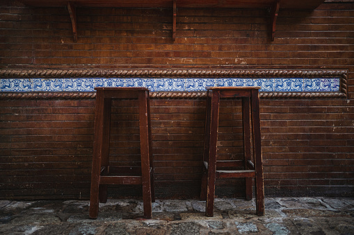 Shot of two high bar wooden stools in front of a wall made of bricks and with a blue spanish design