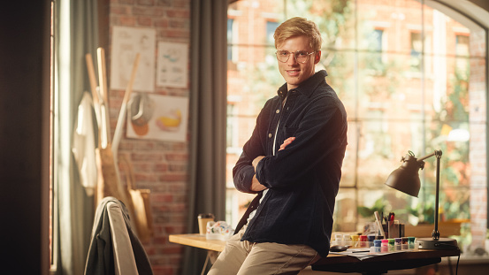 Portrait of Young Caucasian Man with Blonde Hair and Glasses Posing for Camera in Creative Studio. Talented Male Artist Crosses Arms and Looks at the Camera with Smile. Modern Occupation Concept.