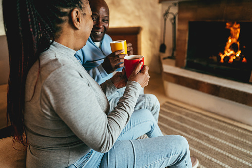 African couple drinking hot chocolate in front of cozy fireplace at home - Winter lifestyle concept - Focus on woman hand