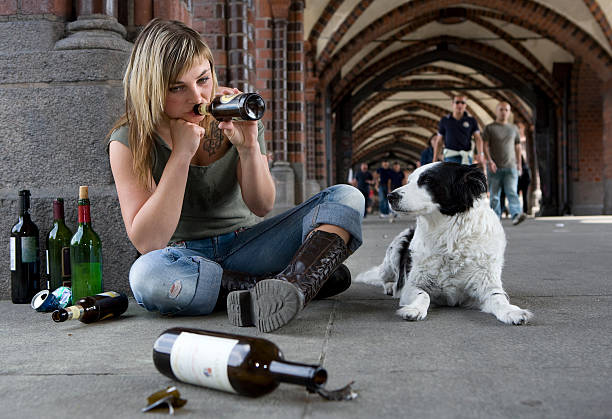 Lonely young woman with dog A young woman in tatter jeans sits on the floor of an ill-conceived BrAcke during the day and drinks alcohol. Her dog lies faithfully next to her and FussgAnger carelessly pass her by. drug addiction tattoos stock pictures, royalty-free photos & images