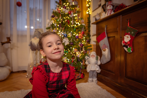 Portrait of a cute little girl sitting at home next to a Christmas tree with lights on Christmas eve.