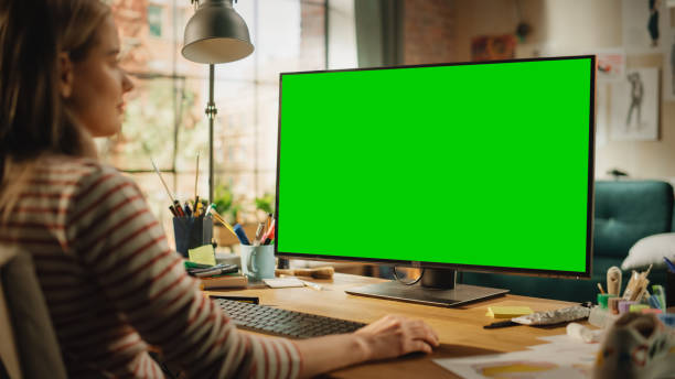 istock Over the Shoulder View of Creative Young Woman Sitting at Her Desk, Using Desktop Computer with Mock-up Green Screen. Female Caucasian Specialist Working on Computer with Chroma Key Display at Home. 1460774508