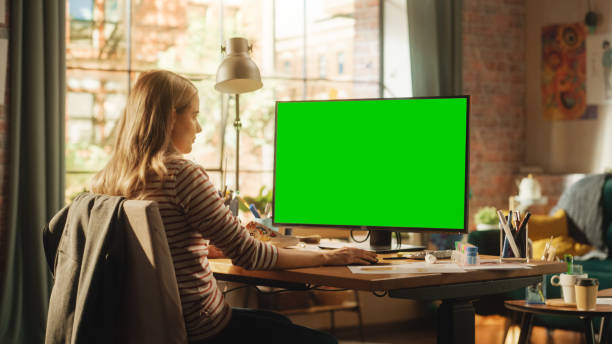 istock Over the Shoulder View: Creative Young Woman Sitting at Her Desk, Using Desktop Computer with Mock-up Green Screen. Female Caucasian Specialist Working on Computer with Chroma Key Display at Home. 1460774582