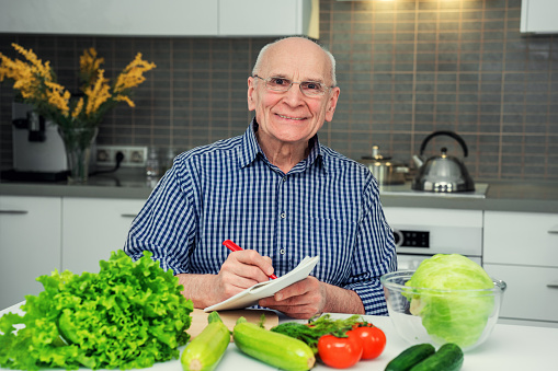 Happy smiling grandfather sitting at table with vegetables and fruits and writing food recipe in notepad. Senior chef ready to vegetarian meal preparation.