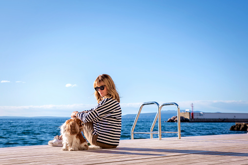 Rear view shot of a woman wearing striped sweater and sunglasses while sitting with her cute dog on jetty by the sea.