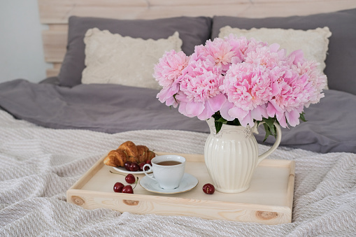 Breakfast in bed. Tray with coffee, croissants and a bouquet of peonies on the bed in the bedroom