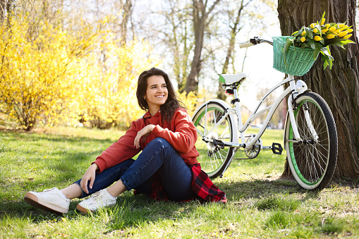 Beautiful woman sitting on green grass near bicycle with bouquet of yellow tulips outdoors. Spring day