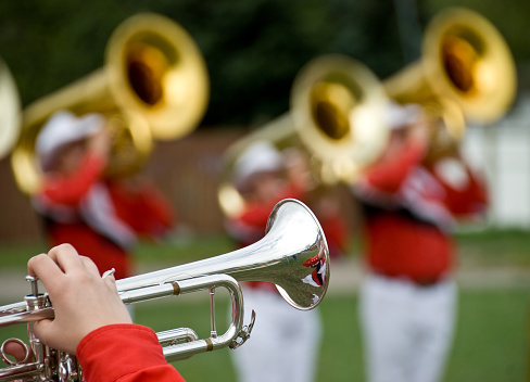 Close view of person playing the trumpet with three out of focus tuba players in the background.