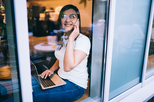 Side view through glass of happy young female student in glasses speaking on cellphone while sitting with laptop near window in cafe