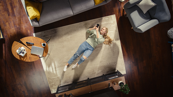 Loft Apartment: Young Woman Lying on a Living Room Floor, Using Smartphone. Girl Relaxes at Home. Freelancer Does Remote Work, E-business, Online Shopping, Social Media Browsing. Top View.
