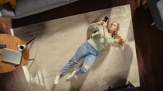 Apartment: Young Woman Lying on a Living Room Floor, Using Smartphone. Girl Relaxes at Home Freelancer Does Remote Work, E-business, Online Shopping, Social Media Browsing. Top View.