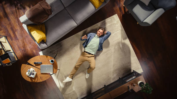 istock Apartment: Handsome Man Lying on a Living Room Floor, Using Smartphone. Guy Relaxes on the Carpet and Looking Up at Camera. Freelance, Remote Work, Online Shopping, Social Media Browsing. Top View. 1460788409