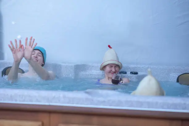 Photo of Cheerful happy company of teenagers enjoys hot bath in hot tub whirlpool on cold winter day. Carefree relaxation in hot tub outdoors of nature. Meeting and chatting with friends
