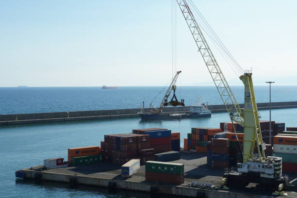 View on movable cargo crane with spreader to handling containers situated in merchant terminal. Genoa, Italy - 08 03 2022: View on movable cargo crane with spreader to handling containers situated in merchant terminal. Near the breakwater is bulk carrier making dredging operation with excavator Terminal Handling Charges stock pictures, royalty-free photos & images