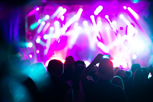 Rear view of people cheering at concert during music festival.Large group of unrecognizable adults with their arms raised.Some of them holding beer cans,some are taping the show with phones.There's a stage and a band performing in background,out of focus.Stage is toned purple blue with lasers and spotlights cutting through smoke and dust.