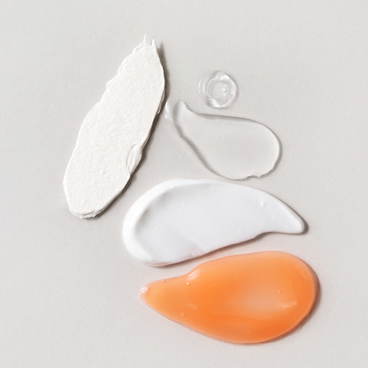 White cream, coral gel or lotion, exfoliating scrub and transparent facial serum swatches. Beauty routine steps. Different skin care products texture. Cosmetic smudge on grey background. Square photo.
