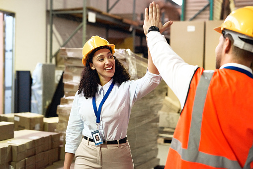 Man, woman and high five in warehouse for logistics, shipping and product transport with safety, ppe and happy. Teamwork, factory staff and motivation with smile, helmet and box for transportation