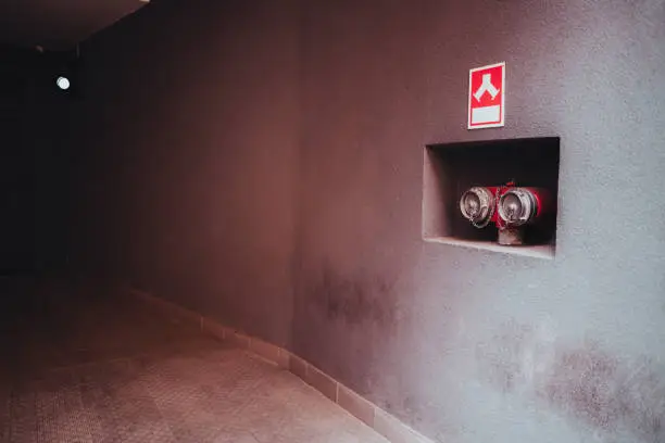 A wide-angle low-key shot of a closed fire fighting doubled hose connector in the recess of the wall with a red fire-pump emblem on the plate above placed in a house or a dark garage entrance
