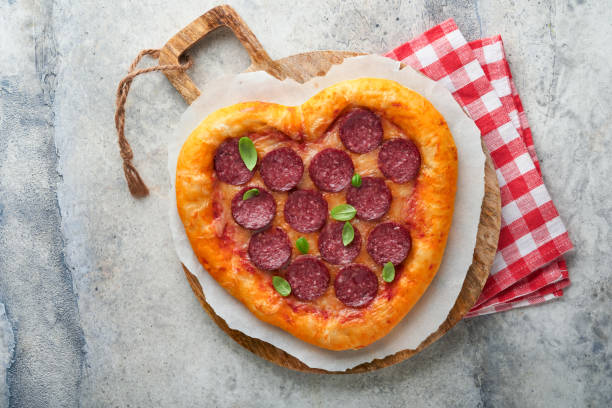 istock Valentines day heart shaped pizza with mozzarella, pepperoni and basil, wine bottle, two wineglass, gift box on light grey background. Idea for romantic dinner Valentines day. Top view. Mock up. 1460825406