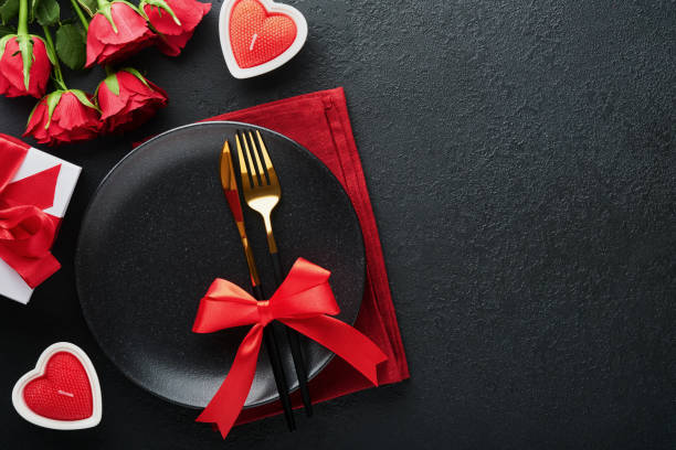 istock Valentines day romantic table setting. Empty closeup of red roses, wine, candles, dinner black plate, knife, fork and decorative silk hearts on black background. Holiday concept. Copy space. Top view 1460825476