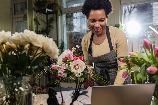 A mature woman is working in her flower shop. She is arranging flower bouquets while talking with a client online via laptop. E-learning.