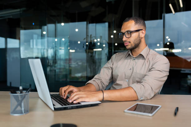 istock Serious and concentrated hispanic programmer working inside office, man in shirt writing codes for software using laptop 1460834931