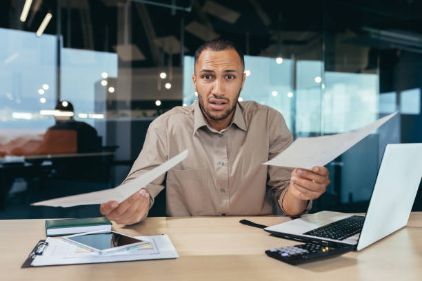 istock Dissatisfied businessman shows documents financial reports to camera, frustrated financier behind paper work looks at camera upset, latin american man working inside office on laptop 1460834985
