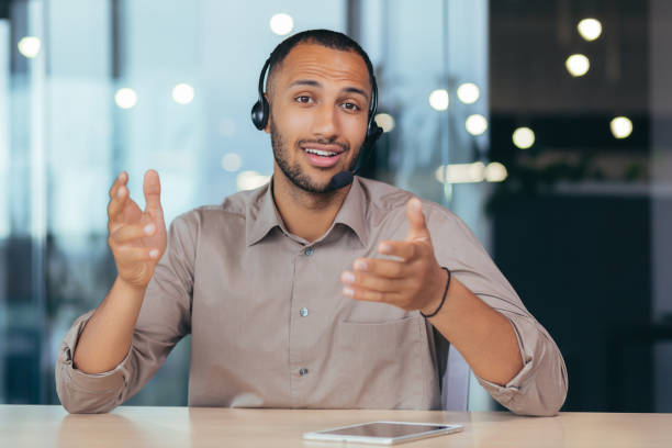 istock Webcam view, happy african american man talking online video call looking at camera using headset, man working inside modern office building tech support and helpline call center 1460835001