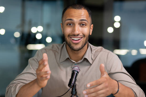 Successful hispanic man inside office looking at camera and talking on video twin inside office, businessman recording audio podcast using professional microphone, webcam view pov
