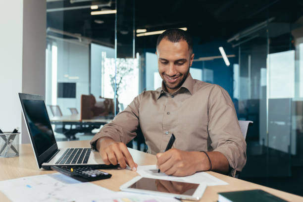 istock Successful african american businessman inside office doing paperwork, man writing information, worker using laptop for work sitting with bills and contracts smiling and satisfied with result 1460835814
