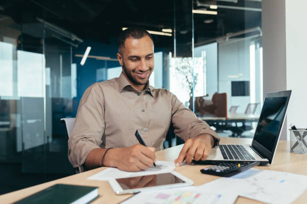 istock Successful african american businessman inside office doing paperwork, man writing information, worker using laptop for work sitting with bills and contracts smiling and satisfied with result 1460836363