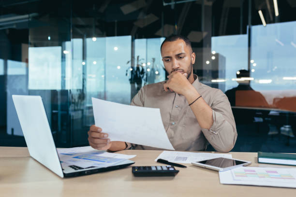 Pensive serious businessman reading financial report, hispanic businessman holding document in hands looking disappointed, working inside modern office with laptop behind paper work. hombre de negocios serio y pensativo leyendo el informe financiero, hombre de negocios hispano sosteniendo un documento en las manos con aspecto decepcionado, trabajando dentro de una oficina moderna con una computadora portátil detrás del papeleo