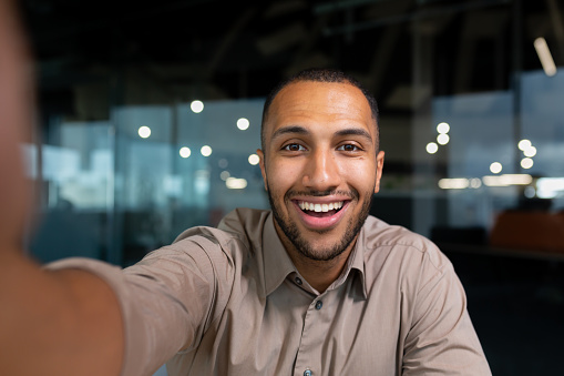 Video ringing, successful businessman looking at smartphone camera talking remotely with colleagues, Hispanic man smiling at work inside office, webcam view pov