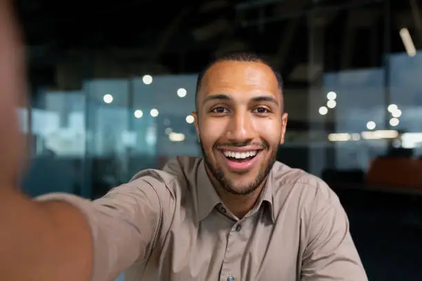 Photo of Video ringing, successful businessman looking at smartphone camera talking remotely with colleagues, Hispanic man smiling at work inside office, webcam view pov