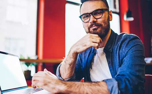 From below of confident bearded male entrepreneur in casual clothing and stylish eyeglasses smiling and looking at camera while watching video on empty screen laptop at cafe