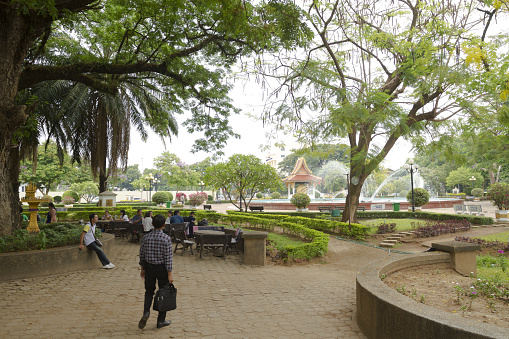 Azalea Park in Vientiane - small public park near Patuxay Monument. A few incidental people are in scene. They are relaxing a tables and benches. A man is walking in foreground. In background is fountain of park